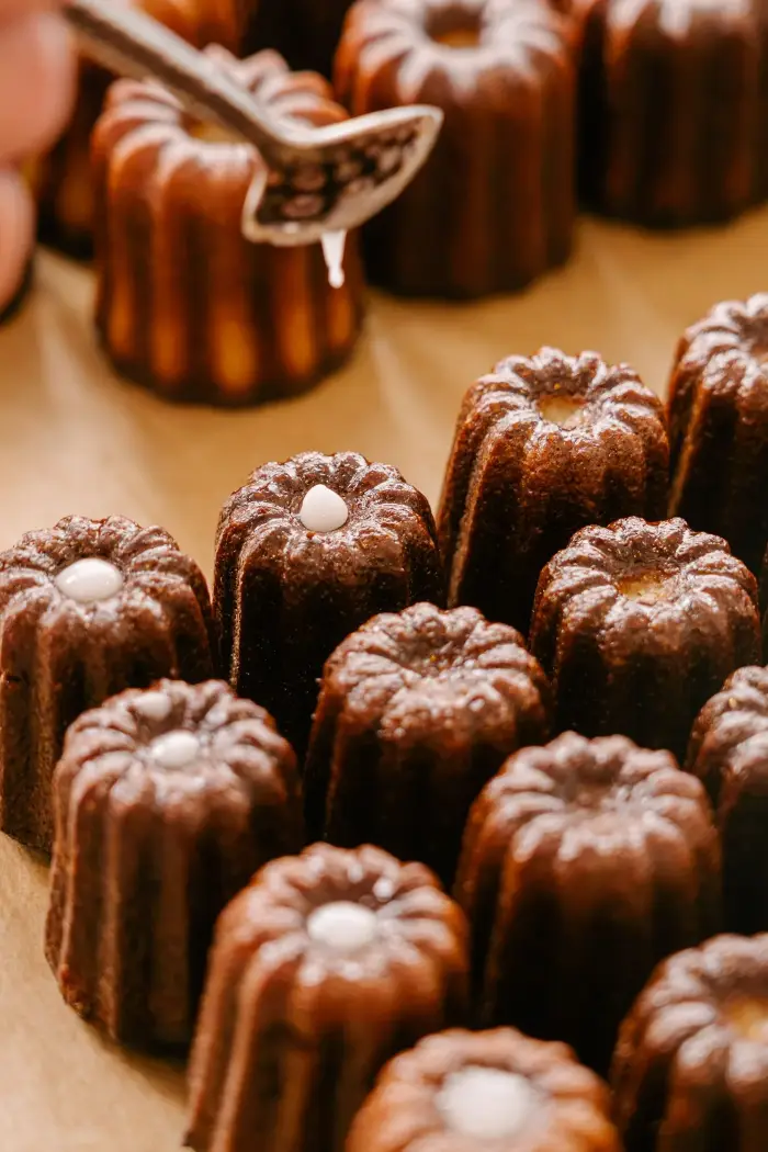 Rangées de cannelés brillants finement caramélisés, chacun garni d'une pointe de crème, préparés à la main chez Coco Cannelés à Paris