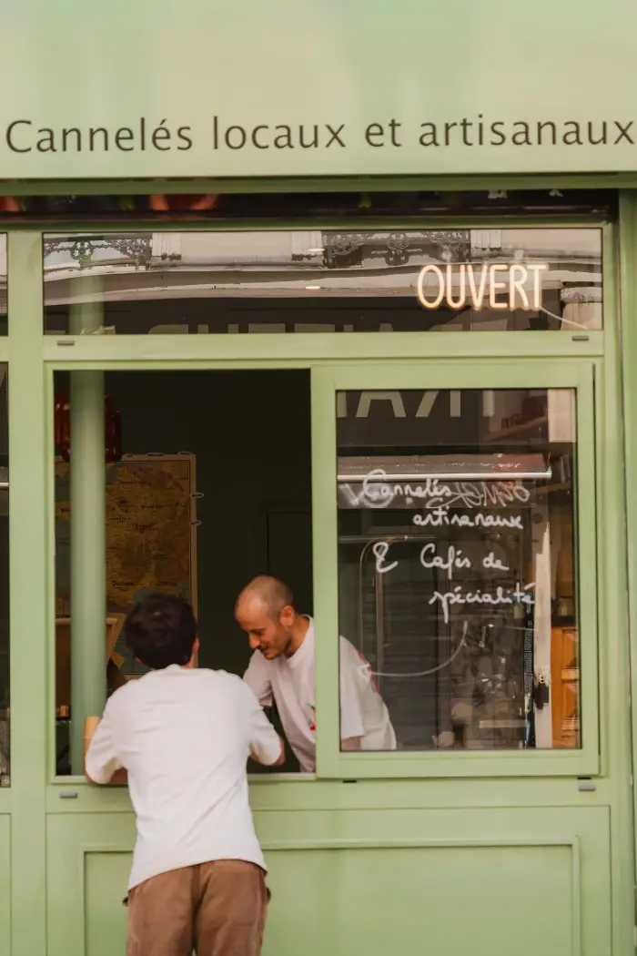 Devanture verte de la boutique Coco Cannelés à Paris, enseigne « Cannelés locaux et artisanaux » et néon Ouvert, un client servi au guichet