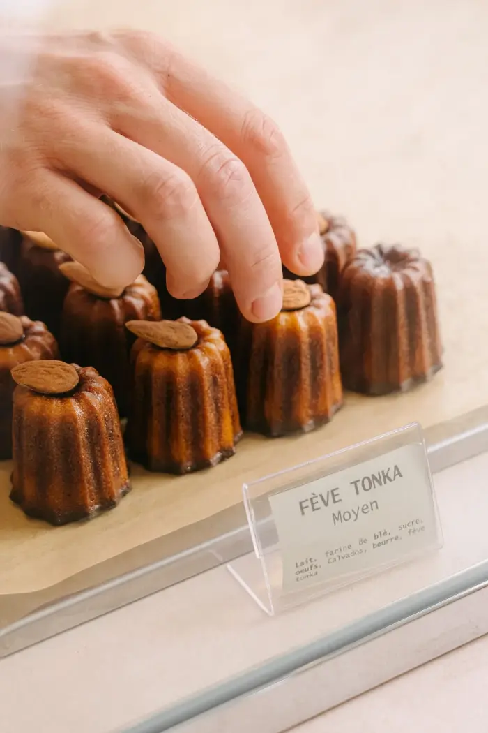 Cannelés à la fève tonka surmontés d'une amande dans la vitrine de la boutique, étiquette « Fève Tonka Moyen » et sa liste d'ingrédients